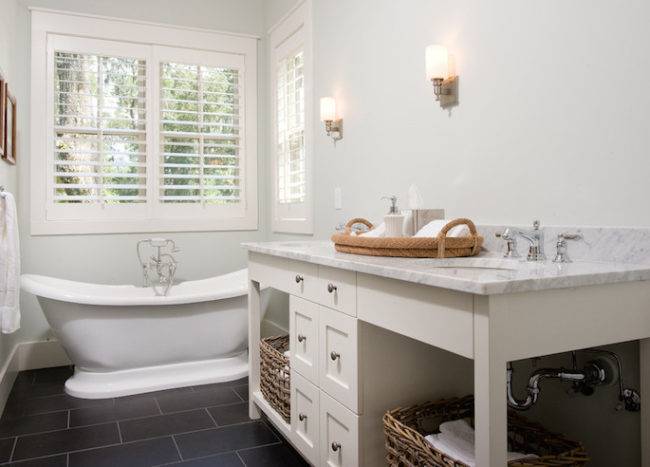 Long cottage bathroom boasts freestanding bathtub placed under windows dressed in plantation shutters across from ivory double washstand with shelves filled with woven baskets topped with carrera marble framing his and her sinks atop black staggered tiled floor.
