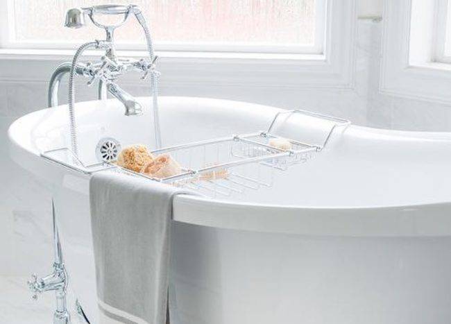 An antique silver claw footed tub sits on marble tiles in front of bay windows and is paired with a polished nickel floor mounted tub filler accenting a polished nickel bath tray.