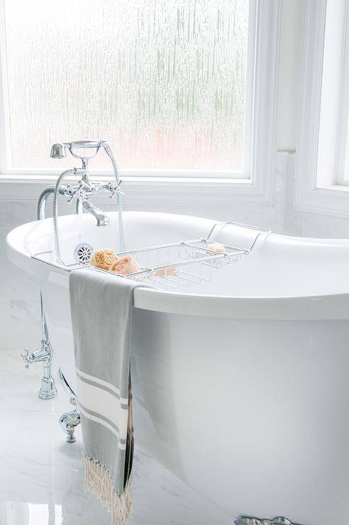 An antique silver claw footed tub sits on marble tiles in front of bay windows and is paired with a polished nickel floor mounted tub filler accenting a polished nickel bath tray.
