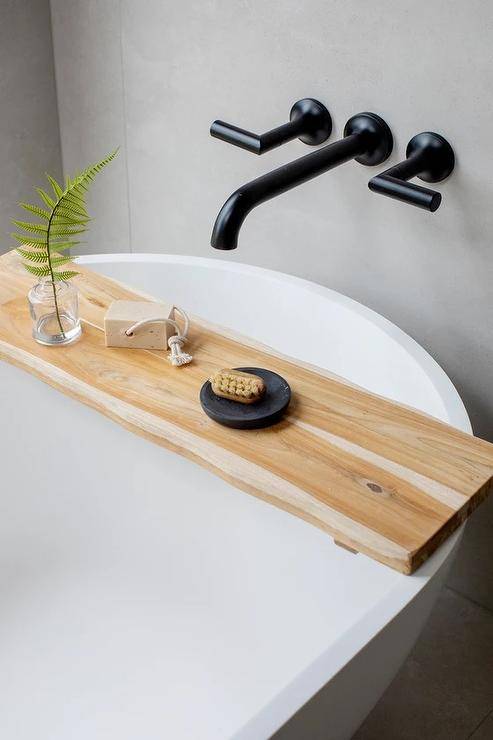 A wooden live edge tray sits on a white bathtub with a matte black wall mount tub filler.