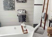 White freestanding oval tub sits in front of a gray shiplap wall on white and black floor tiles. A stained wood tub caddy matches the stained wood towel ladder in front of a white subway tile shower stall.