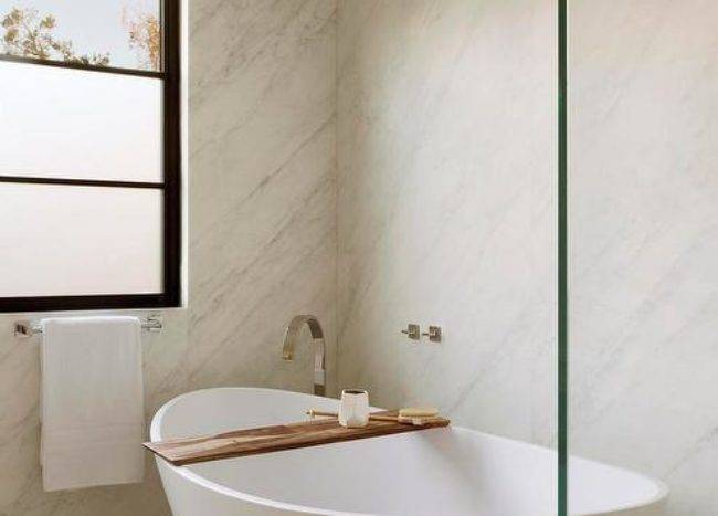 Modern oval tub on white and gold geometric tiles in a custom bathroom boasting a marble slab wall. The bathtub features a teak tray for a contemporary finish.