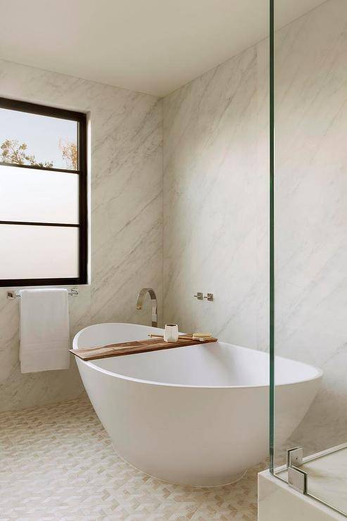Modern oval tub on white and gold geometric tiles in a custom bathroom boasting a marble slab wall. The bathtub features a teak tray for a contemporary finish.