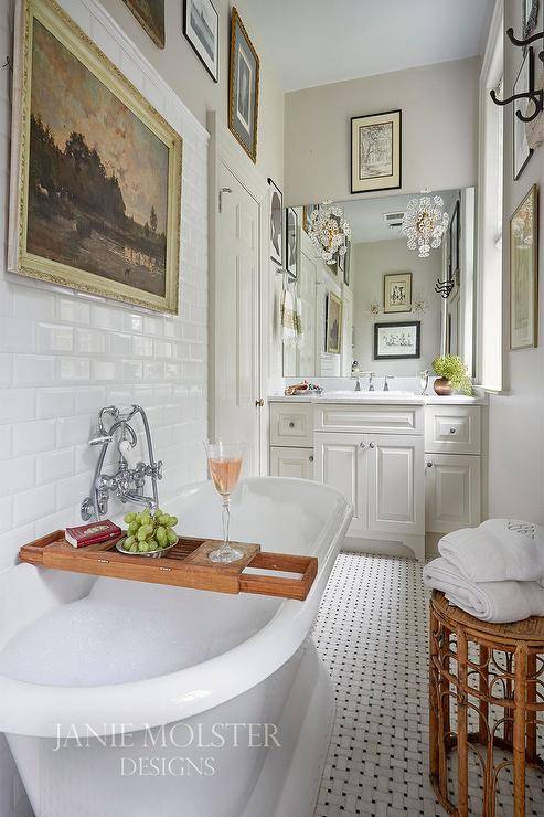 Art hangs from a white beveled subway tiled backsplash over a roll top pedestal bathtub placed on marble basketweave floor tiles and finished with a wooden bath tray and a wall mount polished nickel faucet kit. A round rattan stool sits beside the tub.