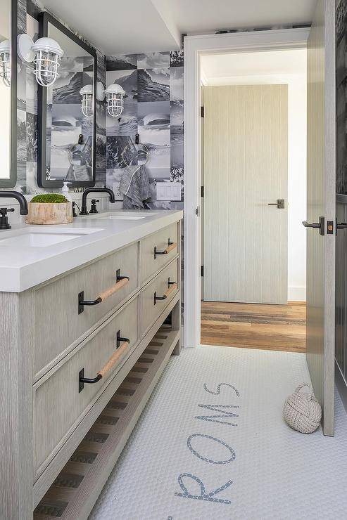 White and gray penny tiles are fixed beneath a gray wash dual washstand boasting a slatted shelf and finished with oil rubbed bronze faucets mounted beneath black Parsons mirrors. The mirrors are lit by white cage sconces mounted against black and white photo collage wallpaper.