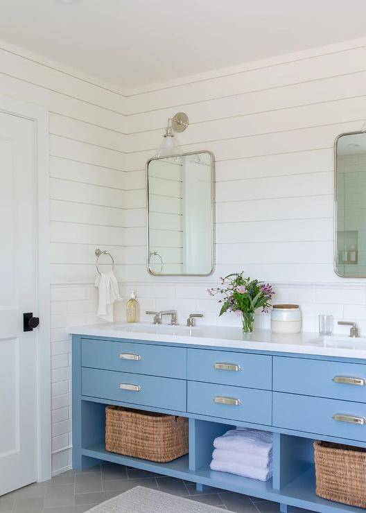 White and blue cottage bathroom boasts a blue dual washstand placed on charcoal gray herringbone floor tiles and fitted with shelves holding woven bins. The washstand is accented with polished nickel cup pulls and polished nickel faucets fixed beneath curved nickel mirrors. The mirror are hung over white staggered backsplash tiles and mounted to white shiplap under glass and nickel conical sconces.