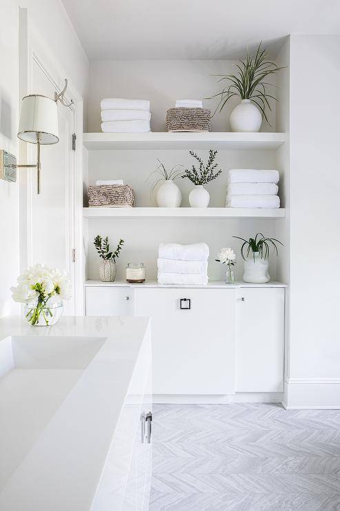 Stacked white floating shelves are fixed in a bathroom nook over white cabinets adorned with square drop pulls and mounted against gray chevron floor tiles.
