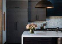 Modern black and white kitchen features a black island fitted with a white quartz waterfall edge countertop lit by a black dome lantern hung from a ceiling finished with wood beams. Sleek black flat front cabinets are adorned with oil rubbed bronze pulls.
