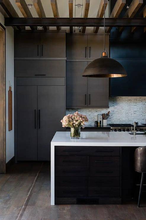 Modern black and white kitchen features a black island fitted with a white quartz waterfall edge countertop lit by a black dome lantern hung from a ceiling finished with wood beams. Sleek black flat front cabinets are adorned with oil rubbed bronze pulls.