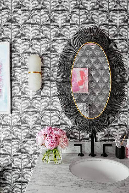 Contemporary bathroom design features a black oval sunburst mirror and glass sconce on gray wallpaper over a light gray sink vanity with a black faucet.