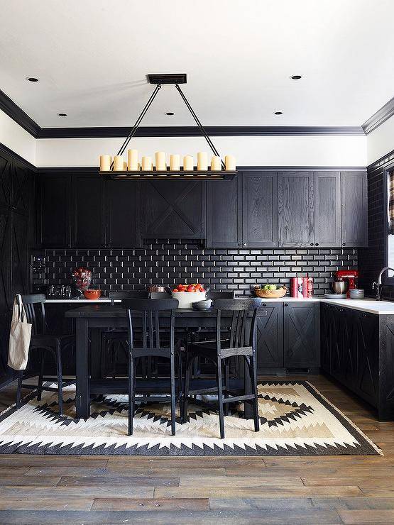 A black pub style island sits on a black and tan rug and is paired with black oak stools. The island is positioned in front of black oak kitchen cabinets contrasted with a white countertop fixed against a black beveled subway tiled backsplash