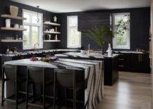 Modern spacious kitchen features a black and white kitchen islands with black leather stools, a black hood over cooktop and light brown floating shelves on black tiles.