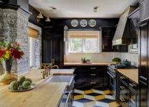 Stylish contemporary black kitchen is accented with a yellow and black painted floor and glossy black cabinets donning wooden countertops and black pulls. A farmhouse sink is matched with an aged brass vintage faucet fixed to a butcher block countertop fixed against a white and black wallpapered wall.