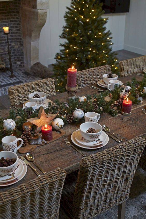 a chic winter table setting with an evergreen, lights and pinecones runner plus wooden stars and ornaments, pillar candles and pinecones in bowls