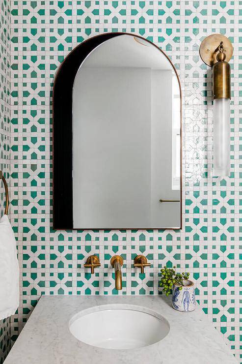 A brass and glass cylinder sconce is fixed to white and green trellis backsplash tiles beside an arched medicine hung over a brass wall mount faucet paired with a marble top bath vanity.