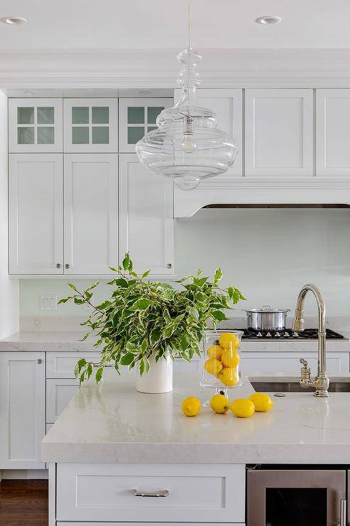 Hudson Valley Lighting Washington Pendant illuminates a white kitchen boasting off-white quartz countertops, white shaker cabinets, and polished nickel knobs. The custom kitchen island features a glass front beverage fridge along with a sink and polished nickel gooseneck faucet. Yellow accents deliver a cheerful, fresh appeal along with lush greenery.