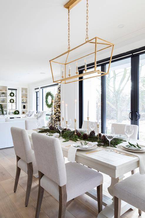 Dining room features a Christmas garland and acorn runner on a long off white dining table with off white fabric dining chairs illuminated by a Darlana rectangular linear lantern.
