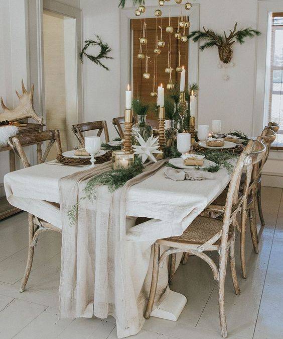a chic and neutral winter tablescape with a neutral runner, greenery, paper stars candles in various candleholders and ornaments hanging over the table