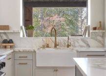 A small picture window is located in a light gray kitchen over an apron sink with an antique brass gooseneck faucet mounted to a marble countertop finishing light gray shaker inset cabinets with brass hardware.