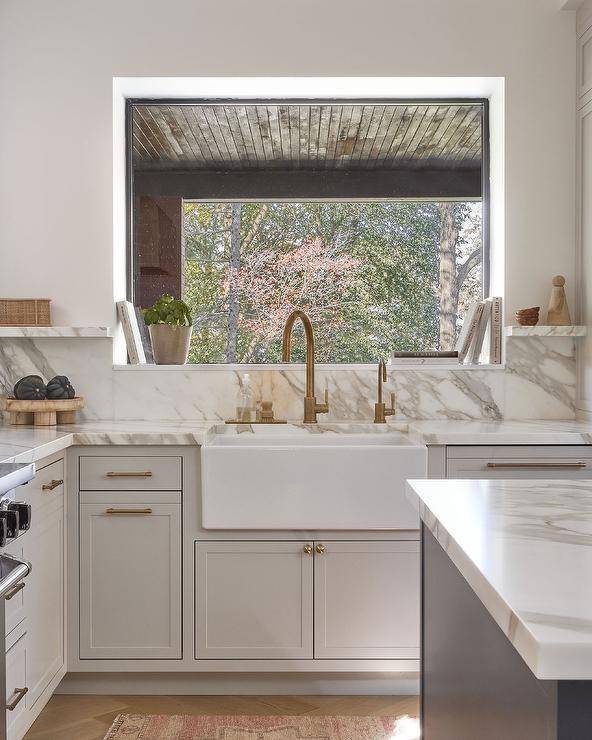 A small picture window is located in a light gray kitchen over an apron sink with an antique brass gooseneck faucet mounted to a marble countertop finishing light gray shaker inset cabinets with brass hardware.