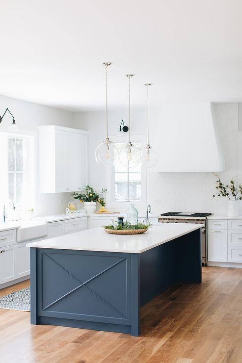 Three glass globe lanterns illuminate a blue x trim island topped with a white quartz countertop surrounded by white shaker cabinets and white backsplash tiles.