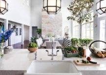 Suzanne Kasler Morris Lanterns over a center kitchen island topped with a white quartz countertop and farmhouse sink. An open floor plan view features lovely dining room decor with a large brick fireplace accent wall.