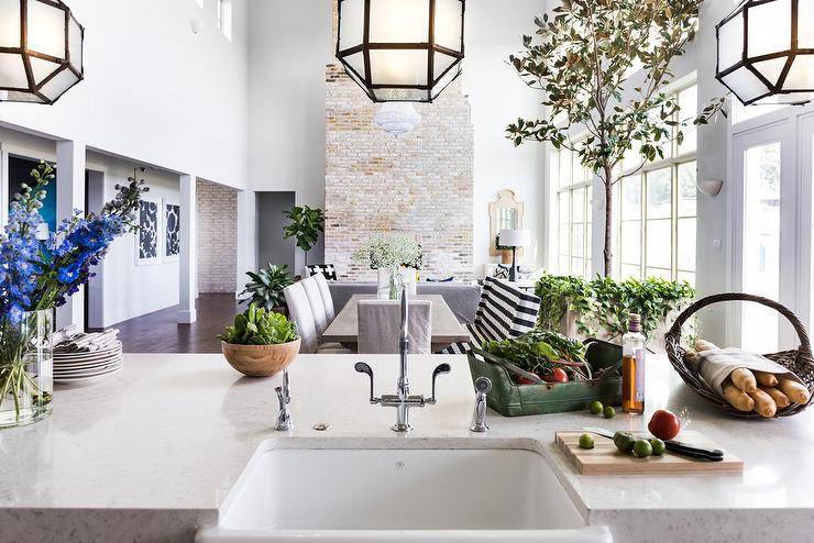 Suzanne Kasler Morris Lanterns over a center kitchen island topped with a white quartz countertop and farmhouse sink. An open floor plan view features lovely dining room decor with a large brick fireplace accent wall.