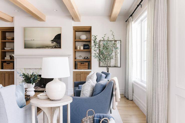 A cream lamp sits atop a round end table placed beside blue upholstered accent chairs located beneath a ceiling finished with wood beams.