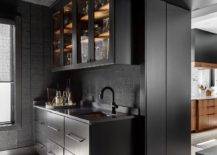 Modern black kitchen pantry is clad in black textured backsplash tiles covering the walls and ceiling. Matte black flat front cabinets donning oil rubbed bronze pulls is topped with a black countertop holding a sink beneath a matte black gooseneck faucet positioned under a glass front cabinet.