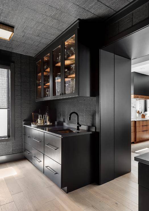 Modern black kitchen pantry is clad in black textured backsplash tiles covering the walls and ceiling. Matte black flat front cabinets donning oil rubbed bronze pulls is topped with a black countertop holding a sink beneath a matte black gooseneck faucet positioned under a glass front cabinet.