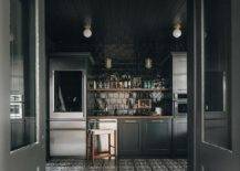 Modern kitchen features a black pantry with a glass door fridge, a wooden floating shelf on glossy black square backsplash tiles, black cabinets, a stainless steel dishwasher, black shiplap ceiling and black and white mosaic pantry floor tiles.