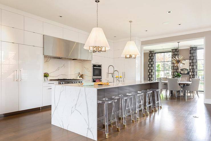 Stunning statuary quartz waterfall island fitted with backless ghost saddle stools finished with brass feet under a set of conical drum light pendants. Wall to wall glossy white lacquer cabinets frame the perimeter kitchen wall showcasing a statuary quartz cooktop backsplash under a long stainless steel vent hood. Wood floors warm up the light cool tones in the kitchen and balance a large, open concept kitchen design.