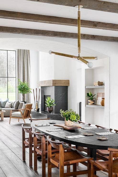 Brown vintage leather chairs an oval black dining table illuminate with modern brass three-light pendants in an open concept dining room design.