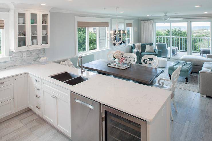 A wine cooler and dishwasher is placed side by side in a kitchen peninsula with a dual sink and gooseneck faucet. Somerset Bay Carmel Arm Chairs frame a dark wood dining table in an open concept kitchen and dining room. A dining bench branches from a kitchen peninsula making good use out of a small space. A white scroll chandelier completes the lighting added as a fixture to the dining room decor.