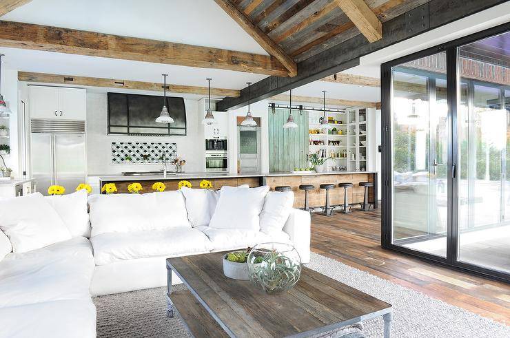 White plush sectional sofa is surely a place to relax and unwind in an open concept, transitional living space. An industrial coffee table on a gray jute area rug under a truss ceiling faces a large glass sliding room door viewing into a backyard patio.