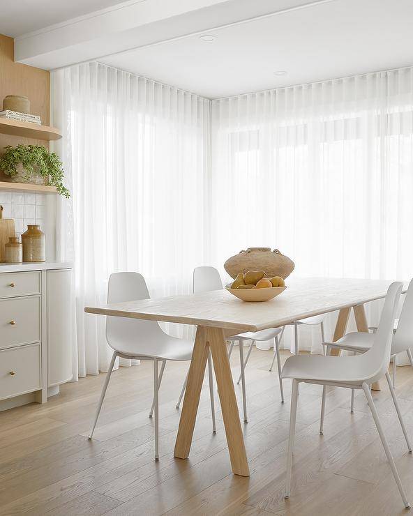 White molded plastic dining chairs sit at a blond dining table in front of windows covered in white pleated curtains.