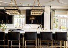 Contemporary black and white kitchen features black cabinetry with brass pulls and gold and black island stools at a marble top island lit by square crystal chandeliers.