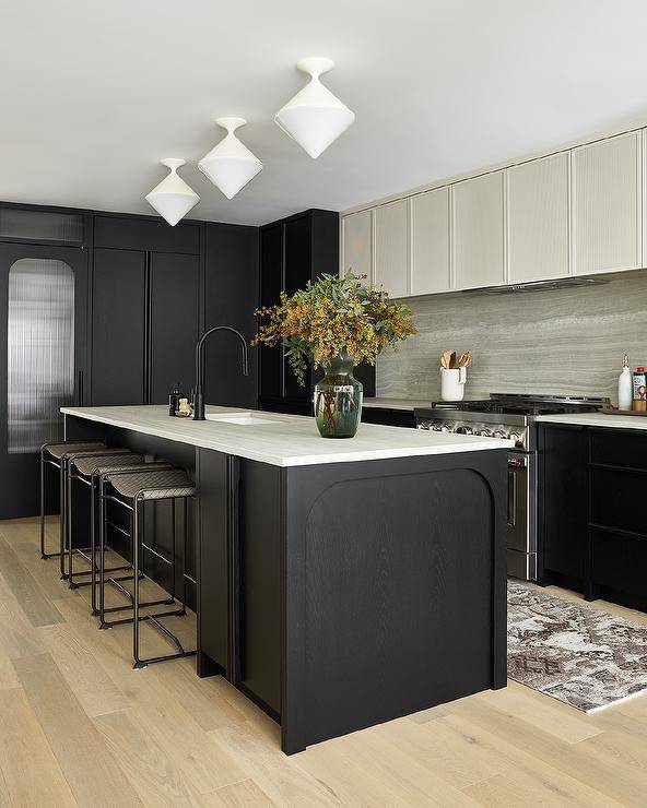 Two-toned ivory and black kitchen features white diamond pendant lights hung over a black oak center island topped with an ivory marble countertop holding a sink with a matte black gooseneck faucet kit front of backless woven gray counter stools. An ivory range hood is fixed over a stainless steel range placed between black cabinets and behind a faded black runner.