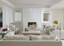 An off-white sofa sits on an off-white rug facing off-white dual coffee tables flanked by side-by-side off-white upholstered chairs lit by a brass floor lamp. A modern white rippled fireplace is flanked by mirrors lit by brass picture lights and hung over white built-in cabinets donning brass pulls.