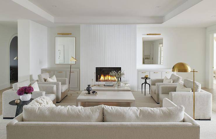 An off-white sofa sits on an off-white rug facing off-white dual coffee tables flanked by side-by-side off-white upholstered chairs lit by a brass floor lamp. A modern white rippled fireplace is flanked by mirrors lit by brass picture lights and hung over white built-in cabinets donning brass pulls.