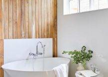 Welcoming bathtub features an asymmetric bathtub placed in front of a teak plank accent wall finished with a polished nickel wall mount tub filler mounted to a white backsplash.