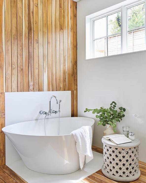 Welcoming bathtub features an asymmetric bathtub placed in front of a teak plank accent wall finished with a polished nickel wall mount tub filler mounted to a white backsplash.