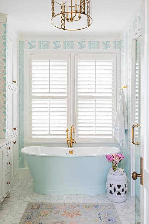 A sky blue freestanding bathtub matched with an aged brass floor mount tub filler sits on marble hexagon floor tiles beneath a window covered in white shutters and framed by Schumacher Hydrangea Drape Wallpaper. A white rope stool is positioned beside the tub.