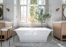 Brushed gold floor mount tub filler paired with a rectangular freestanding tub in a gray tiled bathtub nook. A corner house plant accents the nook with a cozy finish under a window boasting ample natural light.