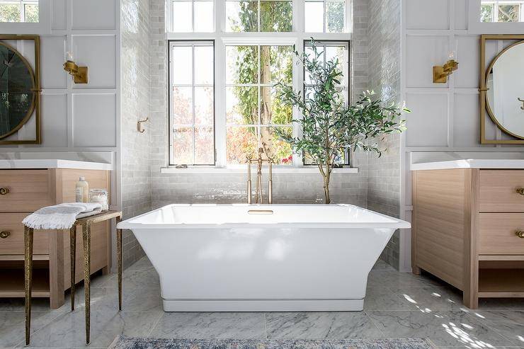 Brushed gold floor mount tub filler paired with a rectangular freestanding tub in a gray tiled bathtub nook. A corner house plant accents the nook with a cozy finish under a window boasting ample natural light.