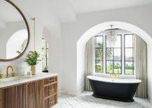 Luxurious bathroom features an arched nook accented with a black and white vintage tub placed on gray and gold marble floor tiles beneath a crystal cascading chandelier, while windows are covered in beige pleated curtains.