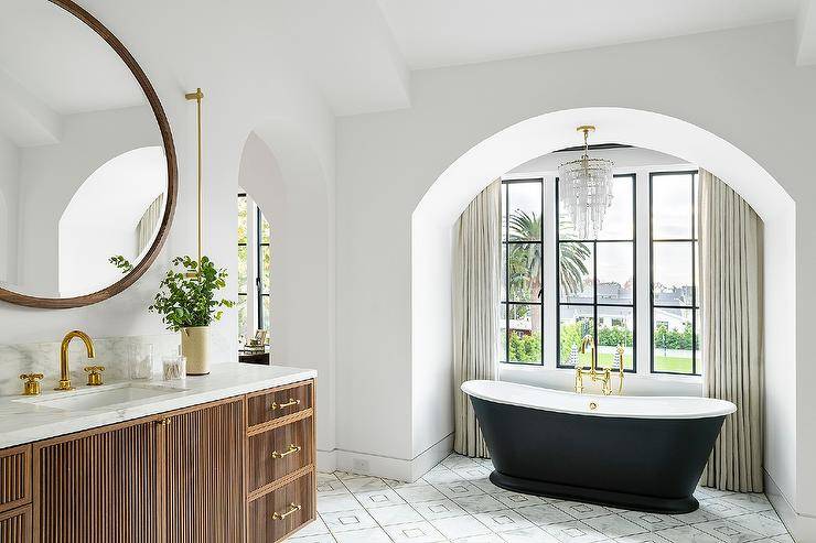 Luxurious bathroom features an arched nook accented with a black and white vintage tub placed on gray and gold marble floor tiles beneath a crystal cascading chandelier, while windows are covered in beige pleated curtains.
