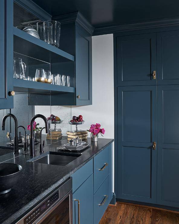 Chic blue pantry cabinets donning brass hardware and a leahter black granite countertop are finished with a stainless steel sink matched with a matte black gooseneck faucet. The faucet is mounted in front of a glass backsplash and beneath stacked blue shelves flanked by blue upper cabinets.