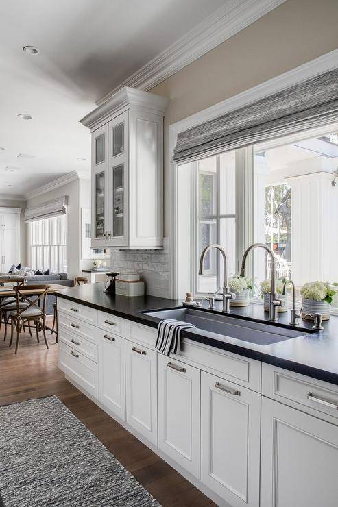 Transitional kitchen with a trough sink and two faucets designed with white cabinets and black leather granite countertops. A window over the trough sink is fitted with a gray roman shade inviting natural light to enhance the kitchens space.