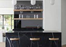 Modern kitchen features a honed black marble bar with wooden shelves, polished black marble countertop and wood and metal tractor stools lit by white pendants.